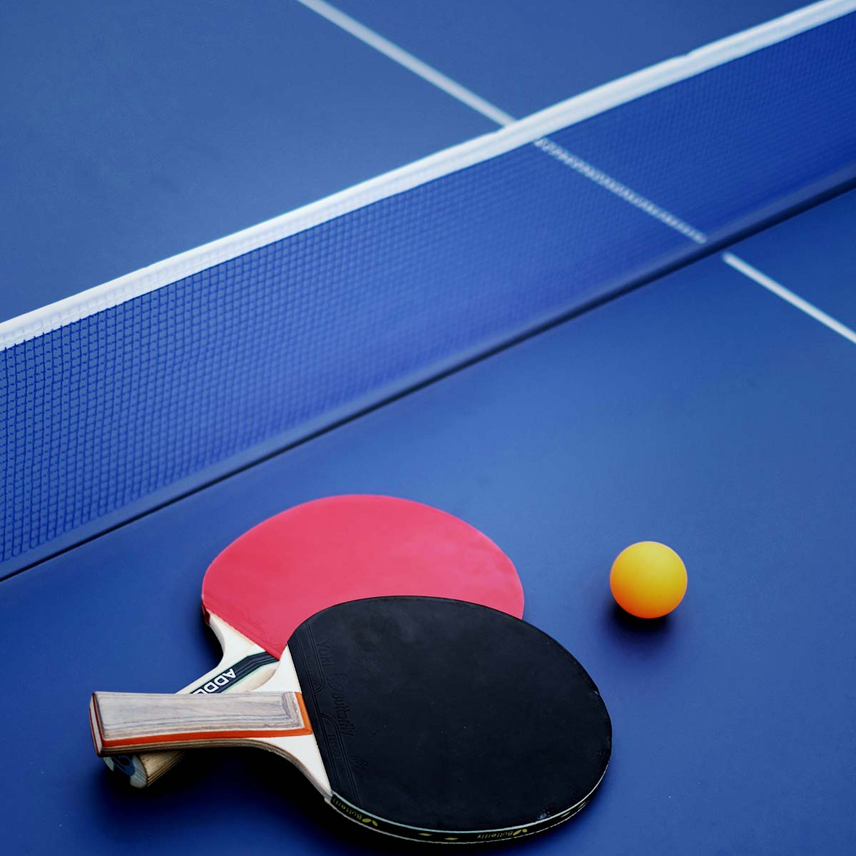 Zwei Tischtennisschläger und ein Ball auf einem blauen Tisch, bereit für ein Spiel.
