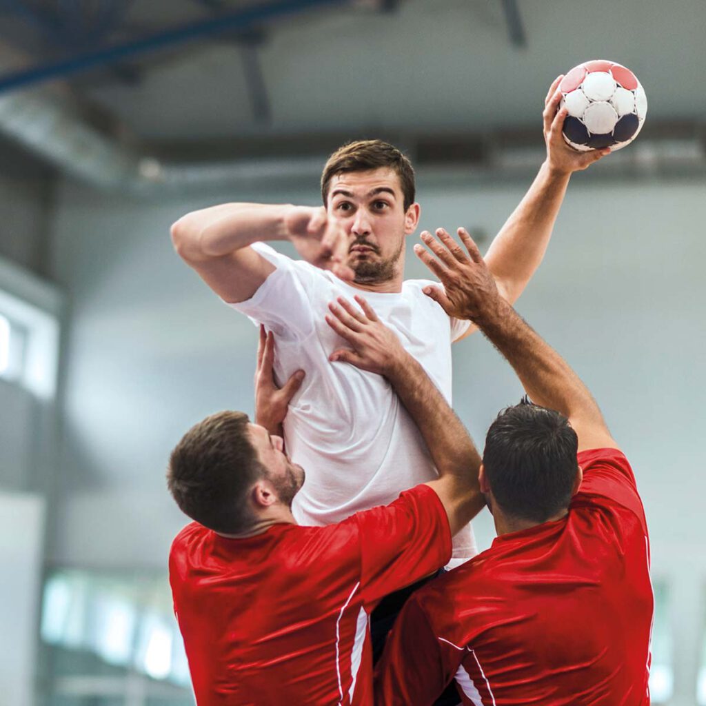 Three men are playing handball in a sports hall. They are active and focused on the game.