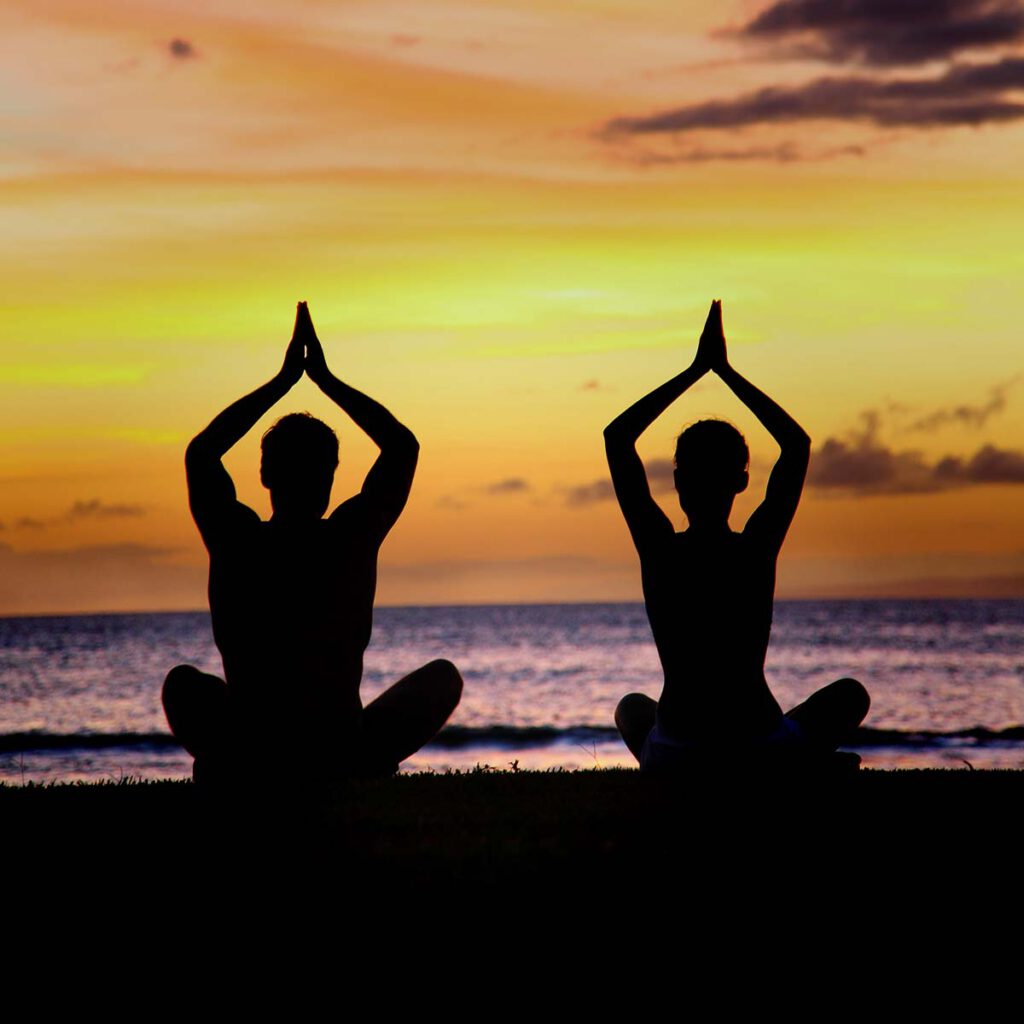 Two people practise yoga at sunset, surrounded by a calm, peaceful atmosphere.